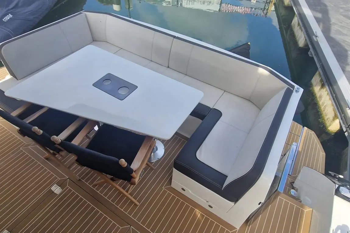 A stylish boat seating area features a light-coloured lounge, a rectangular table with cup holders, and wooden chairs on a teak deck.